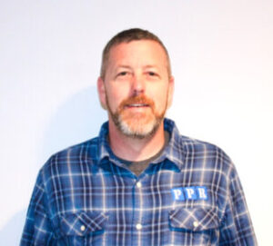 Professional headshot of a man with short hair and facial hair wearing a blue plaid shirt with chest pockets against a white background.