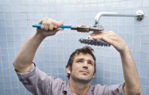 Man in purple shirt inspecting bathroom shower head and pipe fixture against blue tile wall.