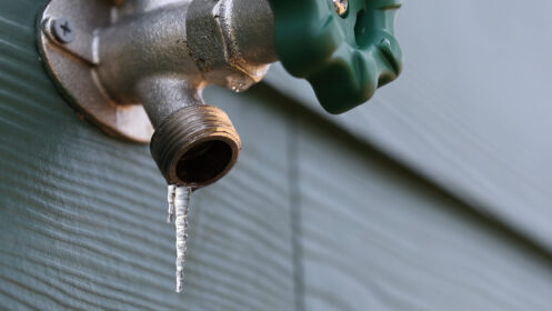 Close-up of a frozen outdoor water faucet with an icicle hanging from the spout, showing frost and ice buildup on the metal fixture.