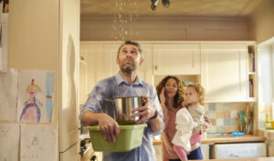 Man holds pot under leaking ceiling while woman and child stand behind him in kitchen during home water damage emergency.