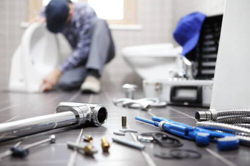 Plumber's tools and equipment on counter with worker in background repairing bathroom fixtures and sink.