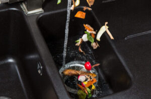 Kitchen sink garbage disposal with water running and food scraps including vegetables and organic waste being disposed of.