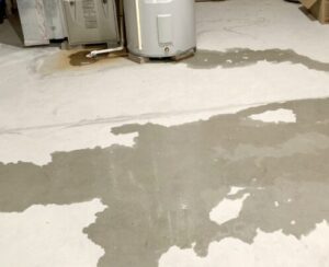 Water puddle spreading across white basement floor near a cylindrical water heater and cardboard boxes.