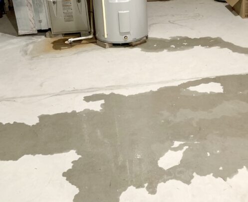 Water puddle spreading across white basement floor near a cylindrical water heater and cardboard boxes.