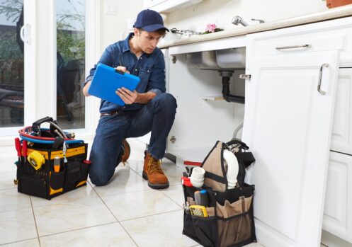 Plumber in blue uniform and cap crouching under kitchen sink with clipboard, toolbags nearby on tile floor.