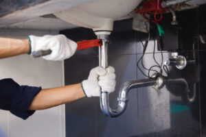 Two workers wearing white gloves installing or repairing pipes and fittings under a sink with a U-shaped trap visible.