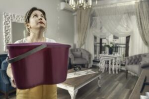 Woman holding burgundy bucket to catch water leak from ceiling in an elegant living room with chandelier and ornate furniture.