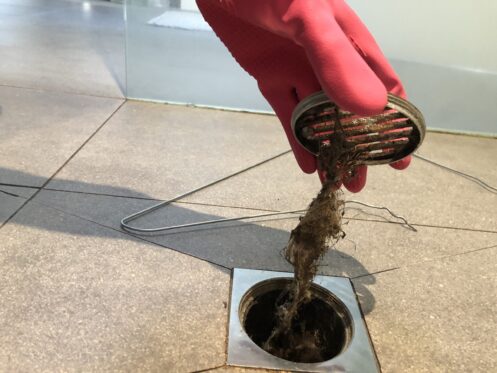 Gloved hand pulling a plumbing snake with debris out of a floor drain pipe during clog removal.