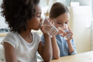 Two young children drinking water from glasses, demonstrating proper hydration and water safety practices indoors.