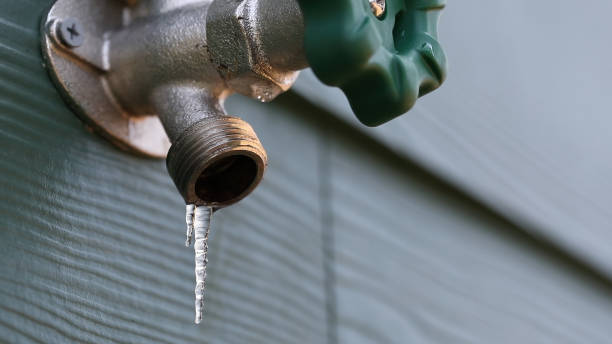 Outdoor faucet covered in frost with an icicle hanging from the spout against a corrugated metal wall.