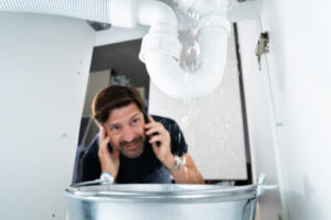 Man on phone looking up at water leaking from ceiling pipe into bucket. Emergency plumbing situation.