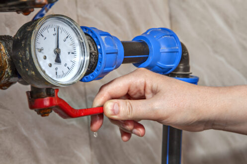 Person's hand adjusting a blue water meter valve with an analog pressure gauge displaying measurement on white dial.