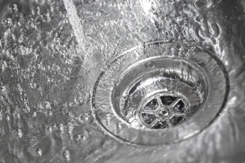 Close-up view of water swirling down a metal sink drain with visible drain cover and splashing water droplets.