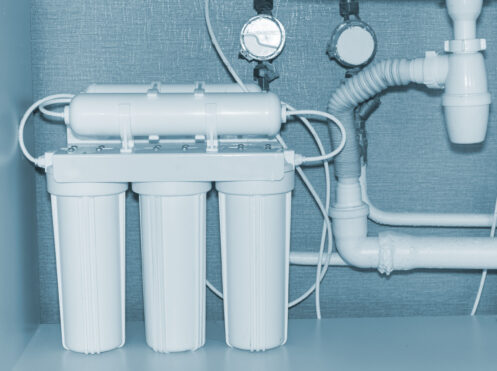 Three-stage white water filtration system installed under a sink with white pipes and pressure gauges mounted on blue wall.