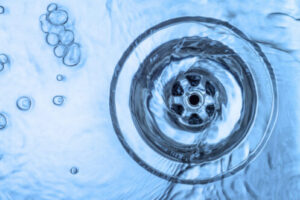 Blue-tinted overhead view of water swirling down a circular drain with bubbles floating on the surface.