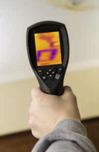 Person holding a thermal imaging camera displaying a heat map with yellow and purple temperature variations on the screen.